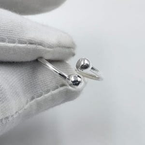Könnte beinhalten: Ein silberfarbener offener Ring mit einem Bypass-Design und zwei polierten kugelförmigen Enden. Das Band ist glatt und metallisch und weist eine reflektierende Oberfläche auf, die das Licht einfängt. Der Ring wird von einer weißen Stoffoberfläche gehalten.