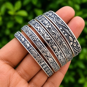 Könnte beinhalten: Ein Set aus fünf silberfarbenen Metallarmreifen mit komplexen Prägemustern. Die Designs umfassen sich wiederholende Blumenmotive, stilisierte Blattformen und eine Reihe kleiner Elefantenfiguren, alles mit einem dunklen, oxidierten Kontrast.