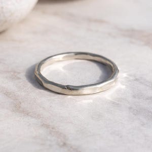 Könnte beinhalten: Ein silberner Ring mit einem strukturierten, facettierten Design. Der Ring ist kreisförmig und scheint handgefertigt zu sein, mit einer leicht unregelmäßigen Oberfläche. Der Ring ist vor einem neutralen Hintergrund platziert, der seinen metallischen Glanz hervorhebt.