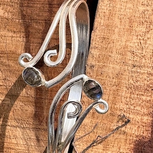 Könnte beinhalten: Ein silberfarbener Ring mit einem verzierten, wirbelnden Design. Der Ring hat zwei ovale, dunkelgraue Edelsteine. Der Ring ist vor einem strukturierten Holzgrund platziert, was seine komplizierten Details hervorhebt.