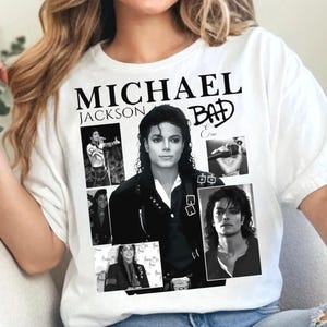 Puede incluir: Camiseta blanca con un collage de imágenes en blanco y negro de Michael Jackson. La camiseta incluye el texto "MICHAEL JACKSON" encima de la palabra "BAD" en una fuente estilizada. También se incluyen imágenes adicionales del artista.