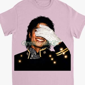 May include: Light pink t-shirt featuring a graphic of a smiling person wearing a black jacket with gold accents and a white glove covered in rhinestones. The person is covering their eyes with the glove. The text "Creatori Co" is visible.