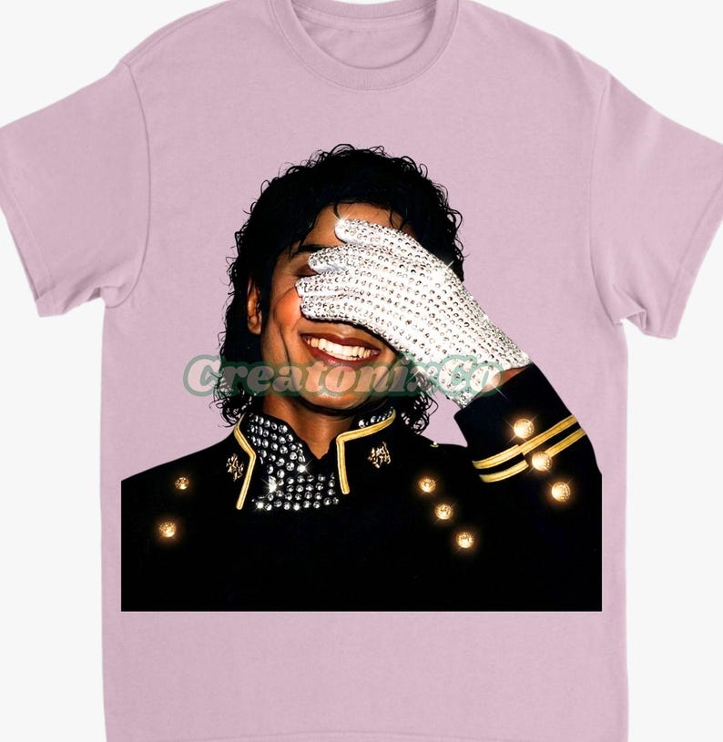 May include: Light pink t-shirt featuring a graphic of a smiling person wearing a black jacket with gold accents and a white glove covered in rhinestones. The person is covering their eyes with the glove. The text "Creatori Co" is visible.