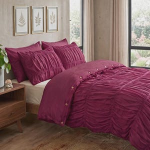 May include: A deep magenta duvet cover set featuring a ruched texture and a row of light-colored buttons along the side. The bedding includes matching pillow shams, all displayed on a bed frame with a cream-colored sheet underneath the duvet.