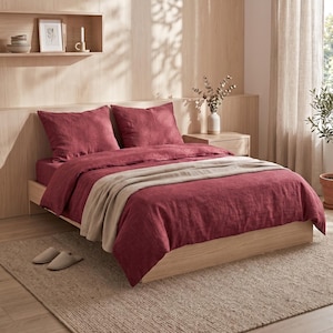 May include: A bed featuring a deep red linen duvet cover and two matching pillow shams. A neutral beige throw blanket is draped across the foot of the bed. The bedding set is displayed on a light-toned wooden bed frame within a bedroom setting.