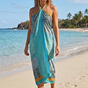 Puede incluir: Un pareo con diseño en tonos azules, turquesas y grises, con un patrón geométrico y floral. El pareo tiene un diseño de chevron turquesa en el centro y un borde dorado. La persona está de pie en una playa de arena.
