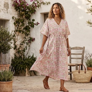 May include: A woman wearing a pink floral print dress with a V-neck and short sleeves. The dress has a drawstring waist and is paired with brown sandals. The background includes a stone wall, plants, and a straw bag.