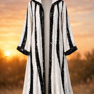 May include: A full-length, hooded coat with vertical black and white stripes. The coat is covered in sequins, with black trim on the hood, sleeves, and hem. The background is a blurred sunset.