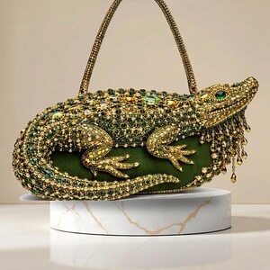 Op de afbeelding: Een ingewikkeld ontworpen handtas in de vorm van een alligator, bedekt met gouden en groene versieringen. De tas heeft een groene basis en een handvat, gepresenteerd op een witte marmeren sokkel. De ogen van de alligator zijn groen.