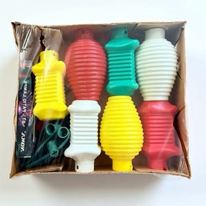 May include: A vintage box of colourful Christmas light bulbs in red, yellow, green, and white. The bulbs have a ribbed design and are arranged in a cardboard box. The box also contains green wire and a small package with text.