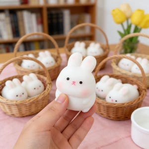 Puede incluir: Una figura de conejito blanco con detalles rosas, sostenida en una mano, con varias figuras de conejito blanco pequeñas en cestas tejidas. Los conejitos tienen ojos negros y mejillas rosas. Las cestas son de material tejido marrón claro.