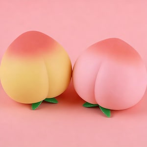 Op de afbeelding: Twee perzikvormige objecten, één geel met een rode bovenkant, de andere roze met een rode bovenkant. Elk heeft een groen blad aan de basis, tegen een roze achtergrond. De objecten zijn ontworpen om op perziken te lijken.