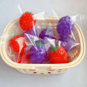 Peut inclure: Un panier jaune clair rempli de fraises et de raisins en plastique, chacun emballé individuellement dans du plastique transparent. Les fraises sont rouges avec des tiges vertes, et les raisins sont violets.