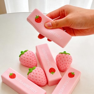 Puede incluir: Gomas de borrar rosas y objetos con forma de fresa. Las gomas tienen la palabra "STRAWBERRY" impresa, junto con un gráfico de fresa roja. Una goma es sostenida por una mano. Las fresas son rosas con tapas verdes.