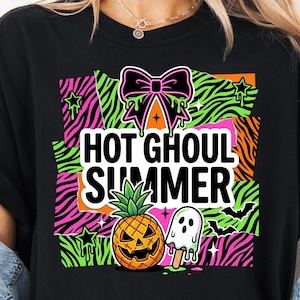 May include: Black t-shirt with a vibrant graphic. The design includes the text "HOT GHOUL SUMMER", a jack-o'-lantern pineapple, a ghost popsicle, and a bow. The background has neon zebra stripes and stars.