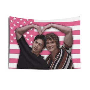 May include: A tapestry featuring two people forming a heart shape with their arms against a pink and white American flag design. One person wears a black shirt, the other a striped shirt.
