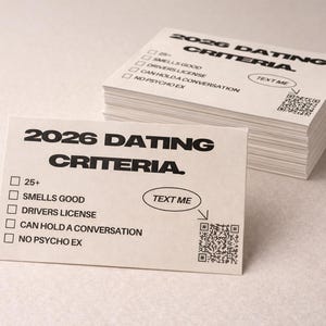 May include: A pile of white cards printed with black text. The cards display "2026 DATING CRITERIA" and include checkboxes for qualities such as "Smells Good" and "Can Hold a Conversation." One card features a "Text Me" prompt and a QR code.