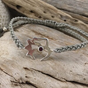 Puede incluir: Un brazalete de cordón trenzado gris con un dije de plata que presenta un diseño estilizado y una piedra preciosa roja.