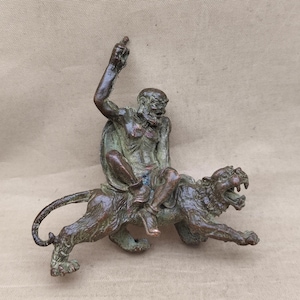 Peut inclure: Sculpture en bronze représentant une figure barbue chevauchant un lion rugissant. La figure, un bras levé, est assise sur le dos du lion. La sculpture a un aspect vieilli, avec des tons verts et bruns.