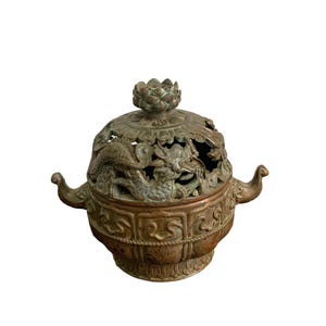 May include: A bronze-toned metal incense burner featuring a removable lid with a lotus-shaped finial and openwork dragon carvings. The bowl has two curved handles on the sides and a base decorated with a repeating geometric pattern and lotus petals.
