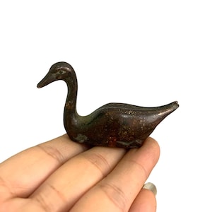 May include: A small, dark brown, metallic figurine shaped like a goose or swan. The object has a smooth, slightly weathered surface with subtle variations in color and a distinct, elongated neck and pointed beak, capturing the bird in a resting pose.