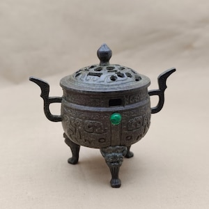 May include: A bronze-coloured incense burner with a decorative lid and three ornate legs. The burner features handles and a green gemstone. The design includes carved patterns and is likely made of metal.