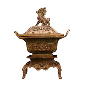 May include: A bronze-toned metal incense burner featuring a rectangular body with ornate relief patterns, two side handles, and a lid topped with a sculpted mythical creature figure. The vessel stands on four curved legs with intricate detailing.