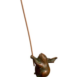 May include: A bronze-toned metal incense holder shaped like a small rabbit standing on its hind legs. The rabbit figure has a weathered, textured finish with hints of green patina and is holding a single, thin, light brown wooden incense stick.