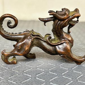 Puede incluir: Figura de color bronce de una criatura mítica, posiblemente un dragón o una bestia similar. La escultura tiene detalles intrincados, incluyendo una cola enroscada, una melena detallada y una boca abierta. La figura está posada en una postura dinámica de caminar.