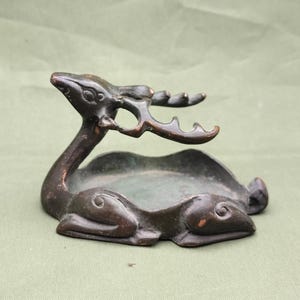 Può includere: Statuetta di cervo color bronzo con un piccolo piatto. Il cervo è in posizione reclinata con la testa e le corna sollevate. Il piatto è di forma ovale e si trova tra il corpo e le corna del cervo. La statuetta ha un aspetto vintage.