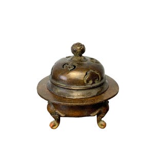 Puede incluir: Un quemador de incienso de metal en tono bronce con una base redondeada, tres patas decorativas y una tapa abovedada extraíble. La tapa presenta intrincados patrones recortados y un pomo central, con un acabado metálico oscuro y desgastado.