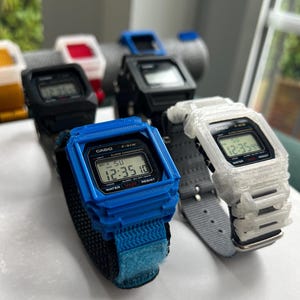 May include: A collection of Casio F-91W digital watches in various colors, including blue, black, white, and yellow. Each watch displays the time and features a rectangular face and a strap. The watches are displayed on a white surface.