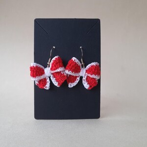 May include: A pair of red and white crochet bow earrings. The bows are red with white trim and are displayed on a black card. The earrings have silver-coloured hooks.