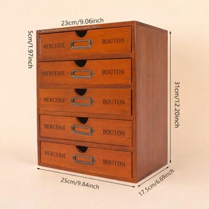 May include: A wooden five-drawer storage cabinet with a warm brown finish. Each drawer is labeled with the words "MERCERIE" and "BOUTON" and has a metal label holder. The cabinet measures 9.84 inches wide, 12.20 inches high, and 6.69 inches deep.
