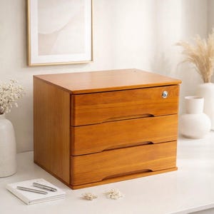 May include: A three-drawer wooden storage cabinet with a warm, medium-brown finish. The cabinet has a lock on the top drawer and recessed drawer pulls. It sits on a white surface, with a notebook and pen nearby.