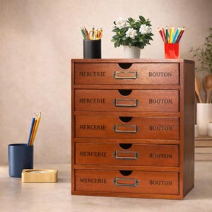 May include: A wooden desk organizer with five drawers, each labeled with the word "MERCERIE" and "BOUTON". The organizer has brass-toned label holders and is filled with pencils, scissors, and a small potted plant. The wood has a warm, reddish-brown finish.