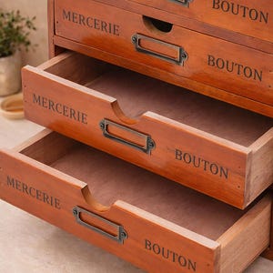 May include: A wooden storage cabinet with multiple drawers, each labeled with the words "MERCERIE" and "BOUTON" in a dark font. The drawers have metal label holders and are stained in a warm brown color.