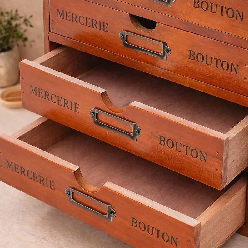 May include: A wooden storage cabinet with multiple drawers, each labeled with the words "MERCERIE" and "BOUTON" in a dark font. The drawers have metal label holders and are stained in a warm brown color.