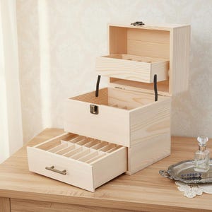 May include: A three-tiered wooden jewelry box with a natural finish. The top section has a lift-up lid, and the middle and bottom sections have pull-out drawers. The bottom drawer has multiple compartments. The box is on a wooden table.