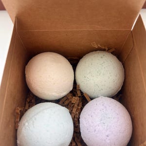 Puede incluir: Un juego de cuatro bombas de baño de colores pastel en una caja de cartón marrón. Las bombas de baño son redondas y tienen una superficie texturizada. Los colores incluyen rosa claro, verde, azul y lavanda. La caja está llena de papel marrón.
