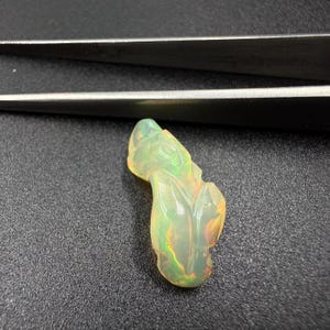 Könnte beinhalten: Ein durchscheinender, unregelmäßig geformter Opal-Edelstein mit einer glatten, wellenförmigen Oberfläche und internen Blitzen in Grün, Gelb und Orange. Der Stein ist in der Nähe einer silberfarbenen Metallpinzette positioniert.