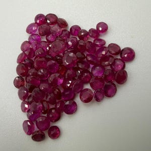 May include: A collection of numerous small, round, faceted rubies with a deep red colour. Each gemstone features a polished surface with multiple geometric facets that reflect light, creating a sparkling appearance across the cluster of loose stones.