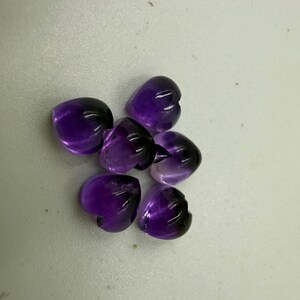 Könnte beinhalten: Eine Ansammlung von fünf kleinen, herzförmigen Perlen aus durchscheinendem violettem Amethyst. Jede Perle hat eine glatte, polierte Oberfläche mit unterschiedlichen Violetttönen, die natürliche Einschlüsse und ein glänzendes Finish aufweisen.