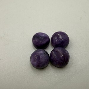 Könnte beinhalten: Vier runde, polierte Cabochons in tiefem Violett mit wirbelnden weißen und dunkelvioletten Einschlüssen. Diese glatten, kuppelförmigen Steine haben eine glänzende Oberfläche und sind in einer quadratischen Formation angeordnet.
