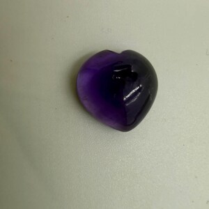 Könnte beinhalten: Ein polierter, herzförmiger Amethyst-Edelstein mit tiefvioletten Farbtönen, durchscheinenden Bereichen und natürlichen Einschlüssen. Die Oberfläche ist glatt und reflektierend und zeigt einen Verlauf von dunklen Violetttönen.