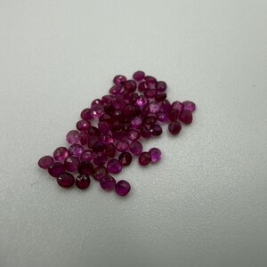 May include: A collection of small, round, faceted loose rubies with a deep magenta and red hue. Each gemstone features a polished surface that reflects light, showcasing a consistent circular cut and vibrant, saturated color throughout the group.