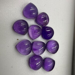 Op de afbeelding: Een verzameling van tien gladde, hartvormige paarse amethist edelstenen. Elke steen heeft een gepolijst, afgerond oppervlak met variërende tinten diep violet en doorschijnende helderheid, losjes gerangschikt op een witte ondergrond.