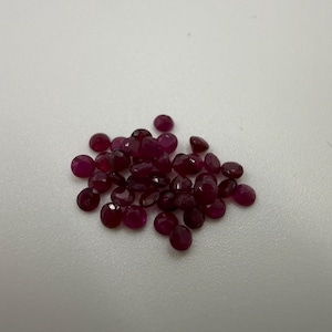 Könnte beinhalten: Eine Ansammlung kleiner, runder, facettierter loser Edelsteine in einer tiefen, leuchtenden magentaroten Farbe. Diese Steine weisen mehrere geschliffene Oberflächen auf, die das Licht reflektieren und ein funkelndes Aussehen erzeugen.