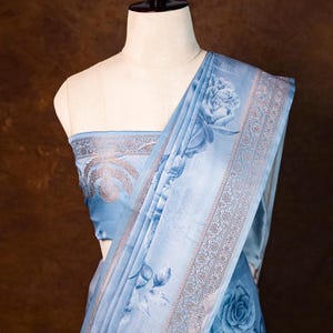 Könnte beinhalten: Ein hellblaues Sari mit einem floralen Rosenmuster. Das Sari ist über einer Schaufensterpuppe drapiert, die das Design des Stoffes zeigt. Der Rand weist ein detailliertes, dekoratives Muster auf. Das Sari ist ein traditionelles Kleidungsstück.