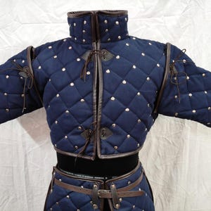 Könnte beinhalten: Ein marineblaues, gestepptes Gambeson mit Rautensteppung und silbernen Nieten. Das Kleidungsstück im mittelalterlichen Stil hat braune Lederbesätze und -bindungen, kurze Ärmel und einen hohen Kragen. Es ist Teil eines Sets, einschließlich eines passenden Gürtels.